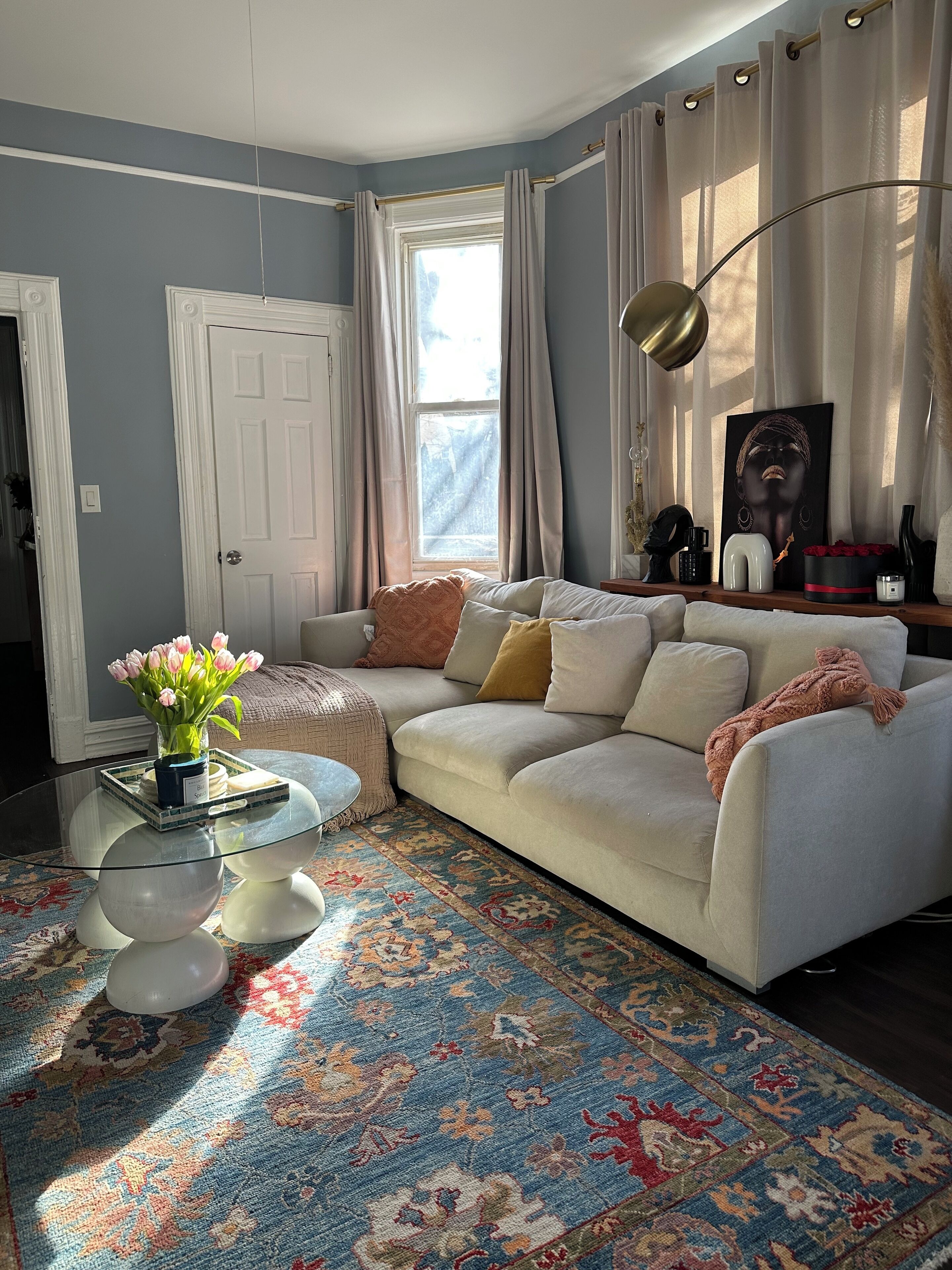 Ein Wohnzimmer mit beiger Couch, einem Glastisch, einem gemusterten Teppich und einem Fenster mit Vorhängen.
