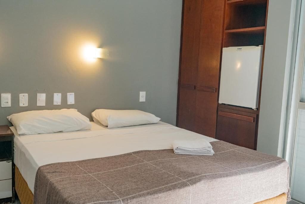 Een hotelkamer met een bed, nachtkastje, wandlamp, kledingkast en koelkast.