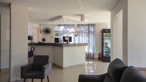 Una sala moderna con un sofá gris, un sillón negro y una zona de cocina con un refrigerador, una máquina de café y un ventilador.