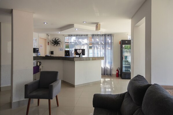 A modern living room with a grey sofa, a black armchair, and a kitchen area with a refrigerator, a coffee machine, and a fan. - HANNA Praia Hotel (Florianópolis)