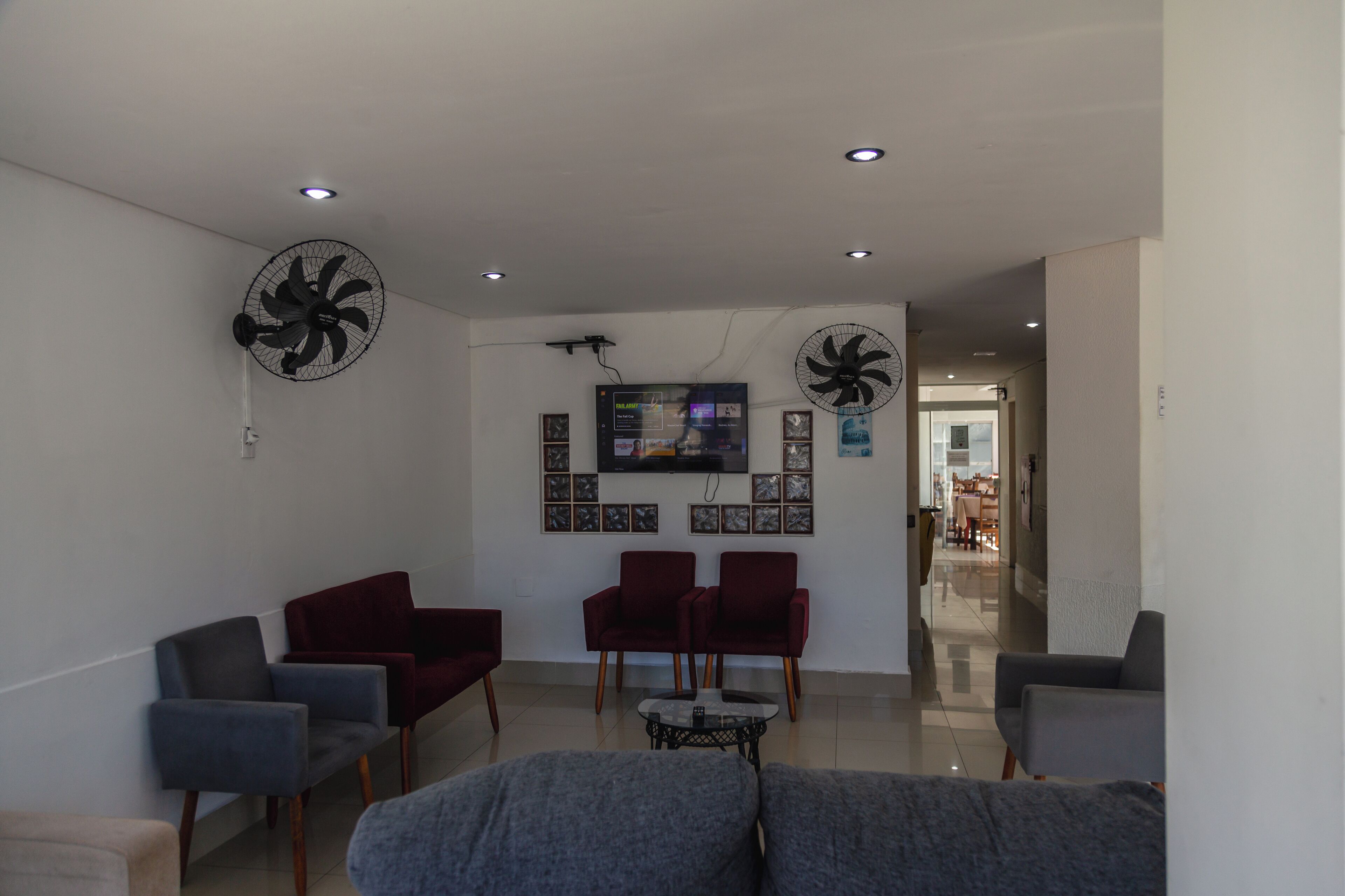 A modern living room with a grey sofa, two red armchairs, and a coffee table. There are two wall-mounted fans and a flat-screen TV on the wall.
