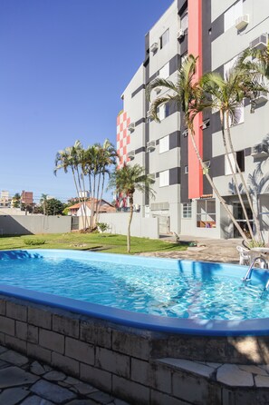 A swimming pool with a clear blue water surface, surrounded by a stone wall. In the background, there is a multi-story building with balconies and a red vertical stripe. Palm trees are visible in the foreground. - HANNA Praia Hotel (Florianópolis)