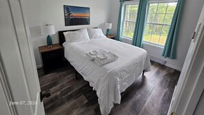 A neatly made bed with white linens, a ceiling fan, a painting of a sunset, and a window with teal curtains. - Acadia Villas!  2A Hunter's Way with EV Charger. (Trenton)
