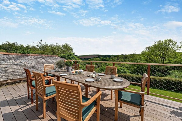 Eine hölzerne Terrasse im Freien mit einem runden Tisch und Stühlen, umgeben von einer Steinmauer und Blick auf grüne Hügel.