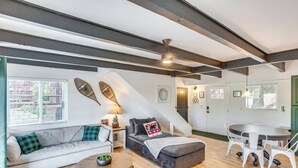 Ein gemütliches Wohnzimmer mit einer Couch, einem Hocker und einem runden Couchtisch. Es gibt einen Essbereich mit Tisch und Stühlen. Im Raum sind Holzbalken sichtbar, und eine Treppe führt zu einer weiteren Ebene.