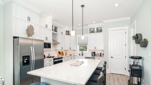 A modern kitchen with white cabinetry, a stainless steel refrigerator, a central island with bar stools, and a coffee machine. - Just steps away - beach, pool, & attractions on the Emerald Isle! New Home.  (Emerald Isle)