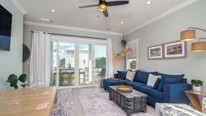A spacious living room with a blue sofa, a wooden coffee table, and a large window offering a view of neighboring houses. - Just steps away - beach, pool, & attractions on the Emerald Isle! New Home.  (Emerald Isle)