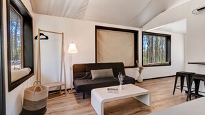 Ein modernes Wohnzimmer mit einem dunklen Sofa, einem weißen Couchtisch und einer Stehlampe. Zwei Fenster bieten Blick auf Bäume, und ein kleiner Beistelltisch trägt eine Vase.
