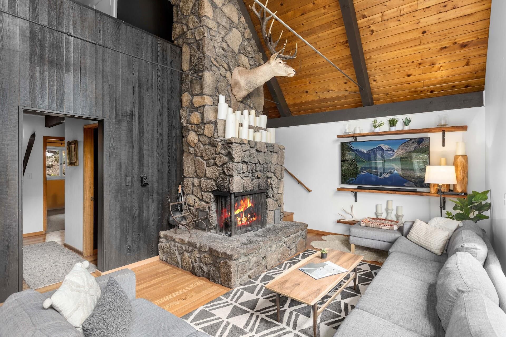 A cozy living room with a stone fireplace, a mounted deer head, and a large painting of a mountain lake.