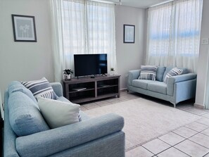 Een woonkamer met twee grijze banken, een flatscreen televisie op een houten dressoir en witte gordijnen.