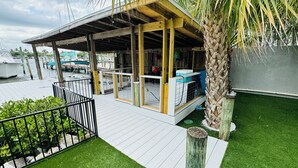 A covered outdoor area with a wooden deck, artificial grass, and a palm tree. - Waterfront Home on the Harbor with a Pool, Boat Dock & Pet Friendly! (Stuart)