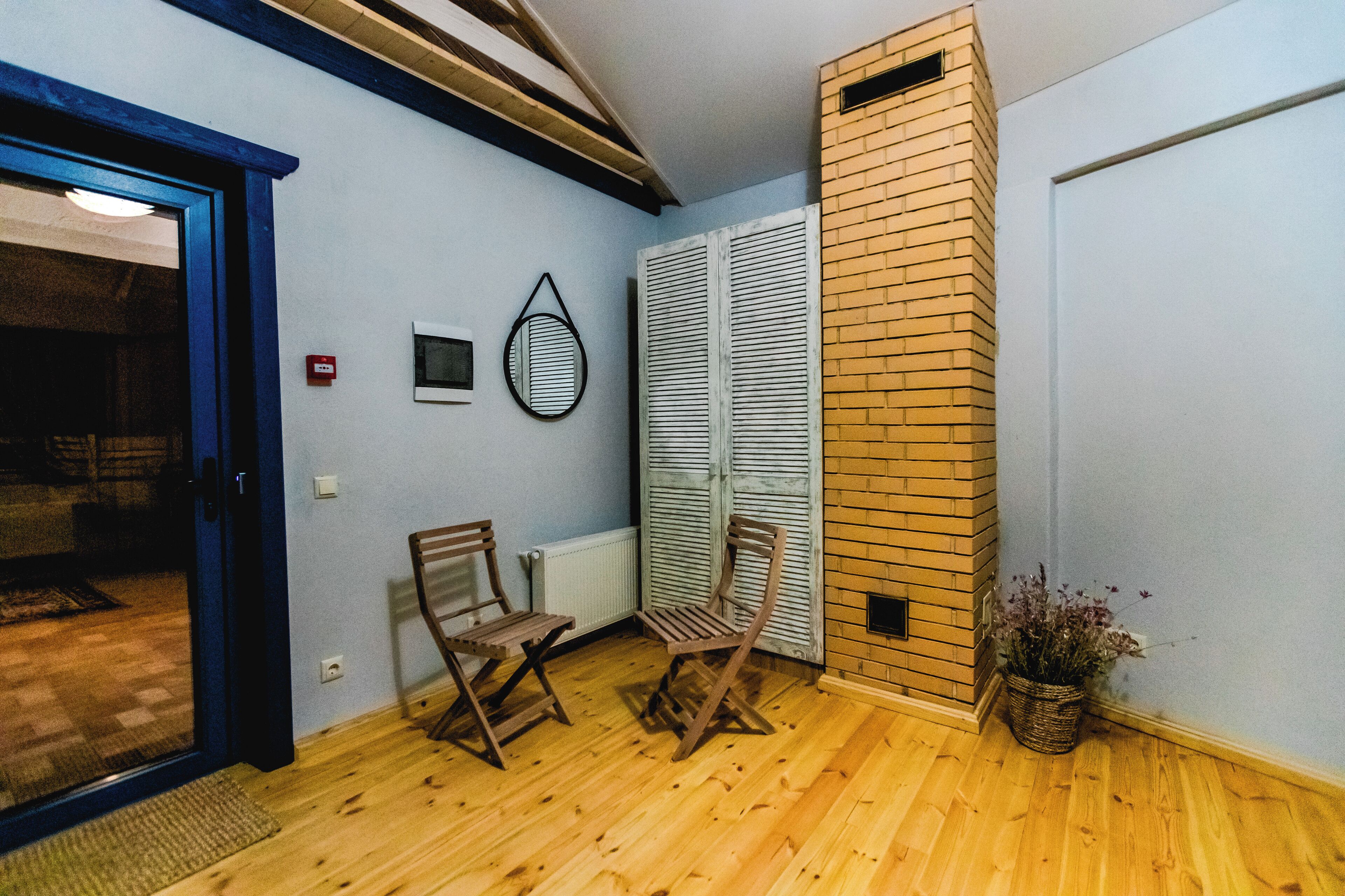 Una habitación con suelo de madera, dos sillas de madera, un radiador blanco, una pequeña planta en maceta y una puerta con marco azul.
