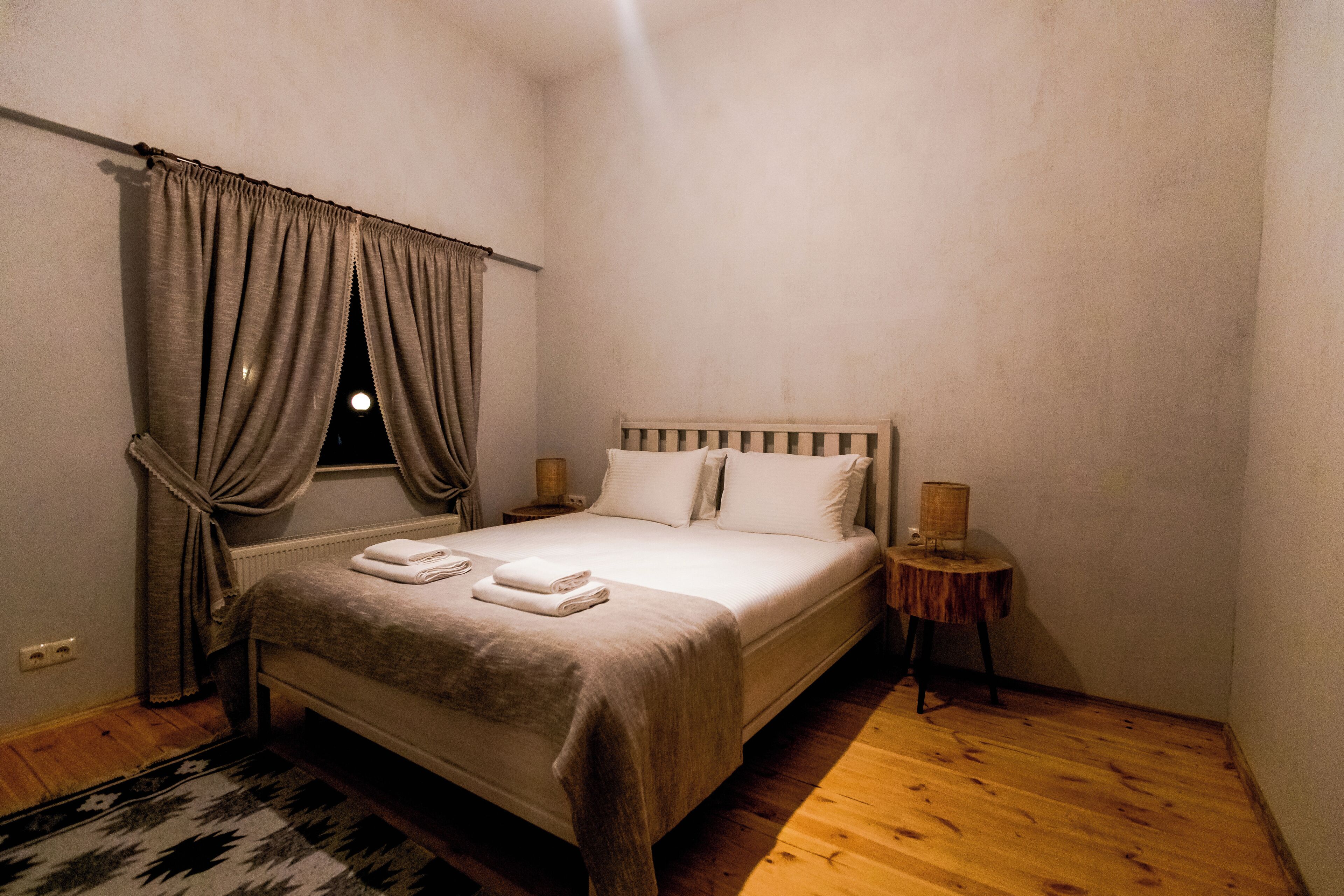 Een slaapkamer met een bed, nachtkastje, lamp en raam met gordijnen.