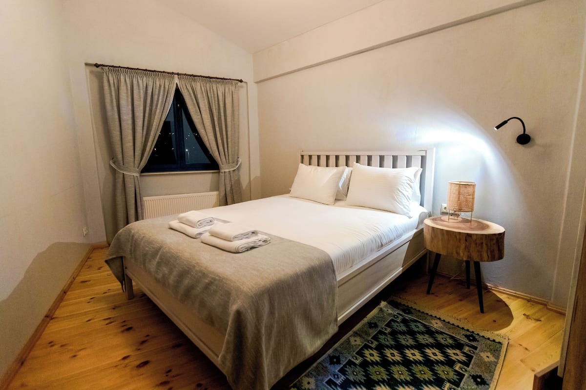 Ein ordentlich bezogenes Bett mit Kopfteil, ein Nachttisch mit Lampe, ein Fenster mit Vorhängen und ein gemustertes Teppich auf Holzboden.