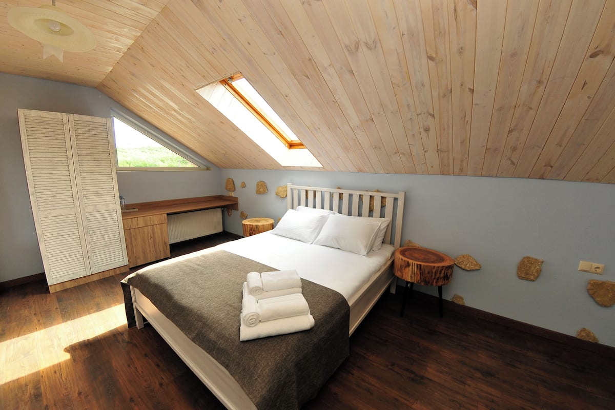 Ein Schlafzimmer mit schrägverlaufender Decke, einem Oberlicht, einem Bett mit grauer Bettdecke, einem Holztisch und einem kleinen runden Tisch.