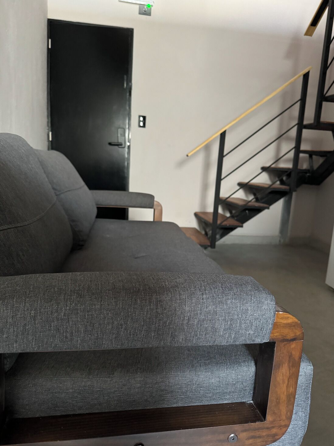 A grey sofa with wooden legs in a room with a staircase and a door in the background.