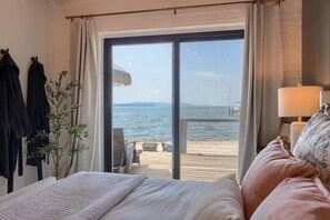 Une chambre avec une grande fenêtre donnant sur une plage, un lit, une table de chevet et une lampe.