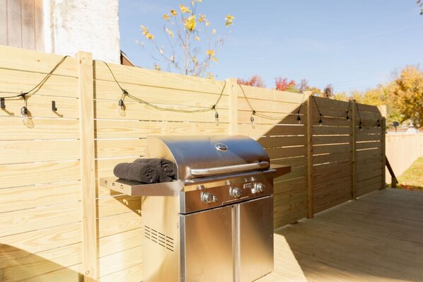 A stainless steel barbecue grill with a towel on it, set against a wooden fence with string lights. - Long Beach Cottage, (romantic couple’s getaway) (Charlestown)