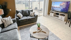A living room with a grey sofa, a coffee table with a plant, a TV on a wooden stand, and a ceiling fan. - Vacation Fantasia - Near Disney (Kissimmee)