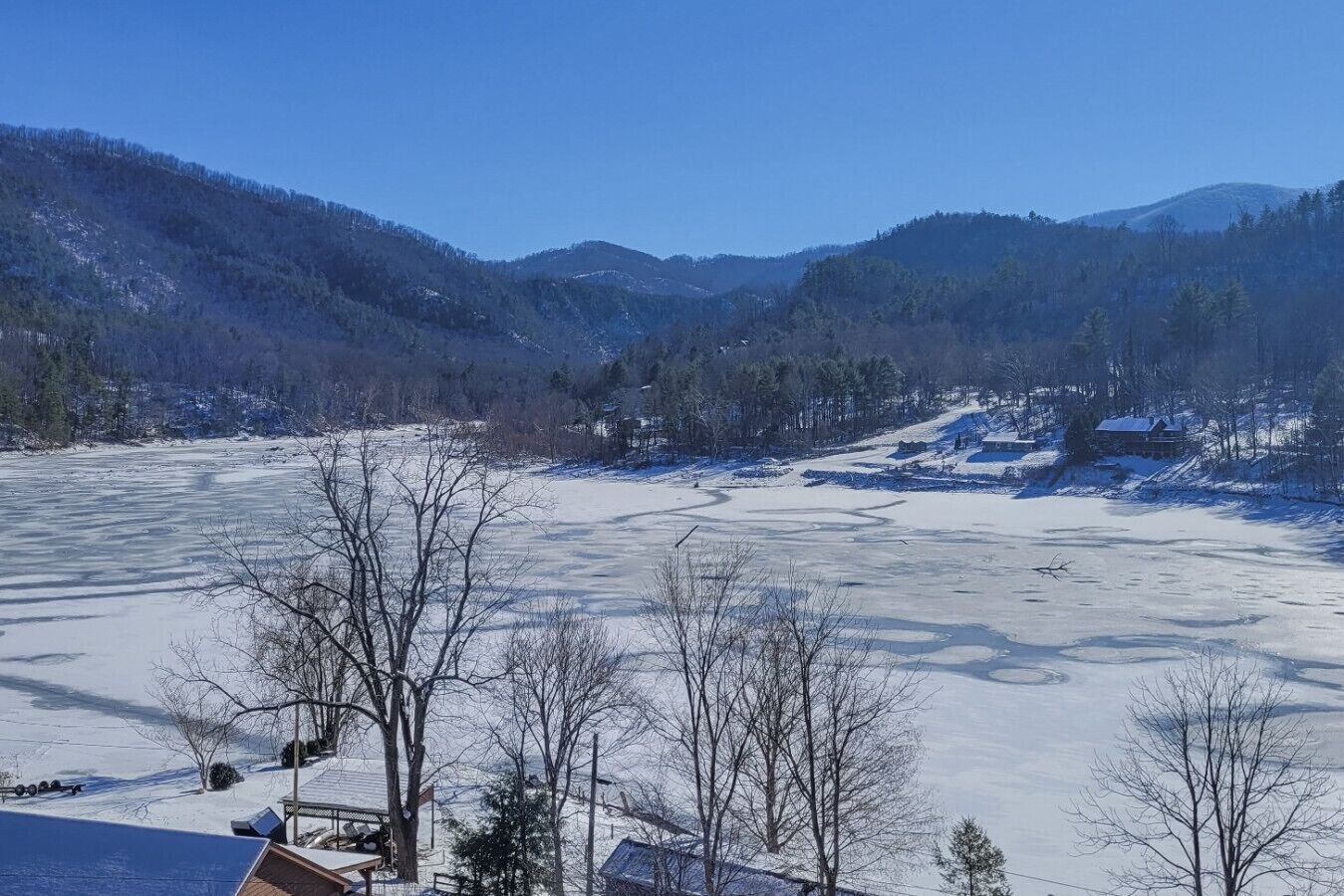 Schneebedeckte Landschaft mit einem zugefrorenen See, Bäumen und Häusern im Hintergrund.