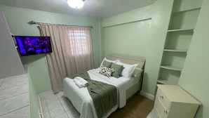 Ein Schlafzimmer mit Bett, Fernseher, Fenster mit Vorhängen und einem kleinen Schrank.