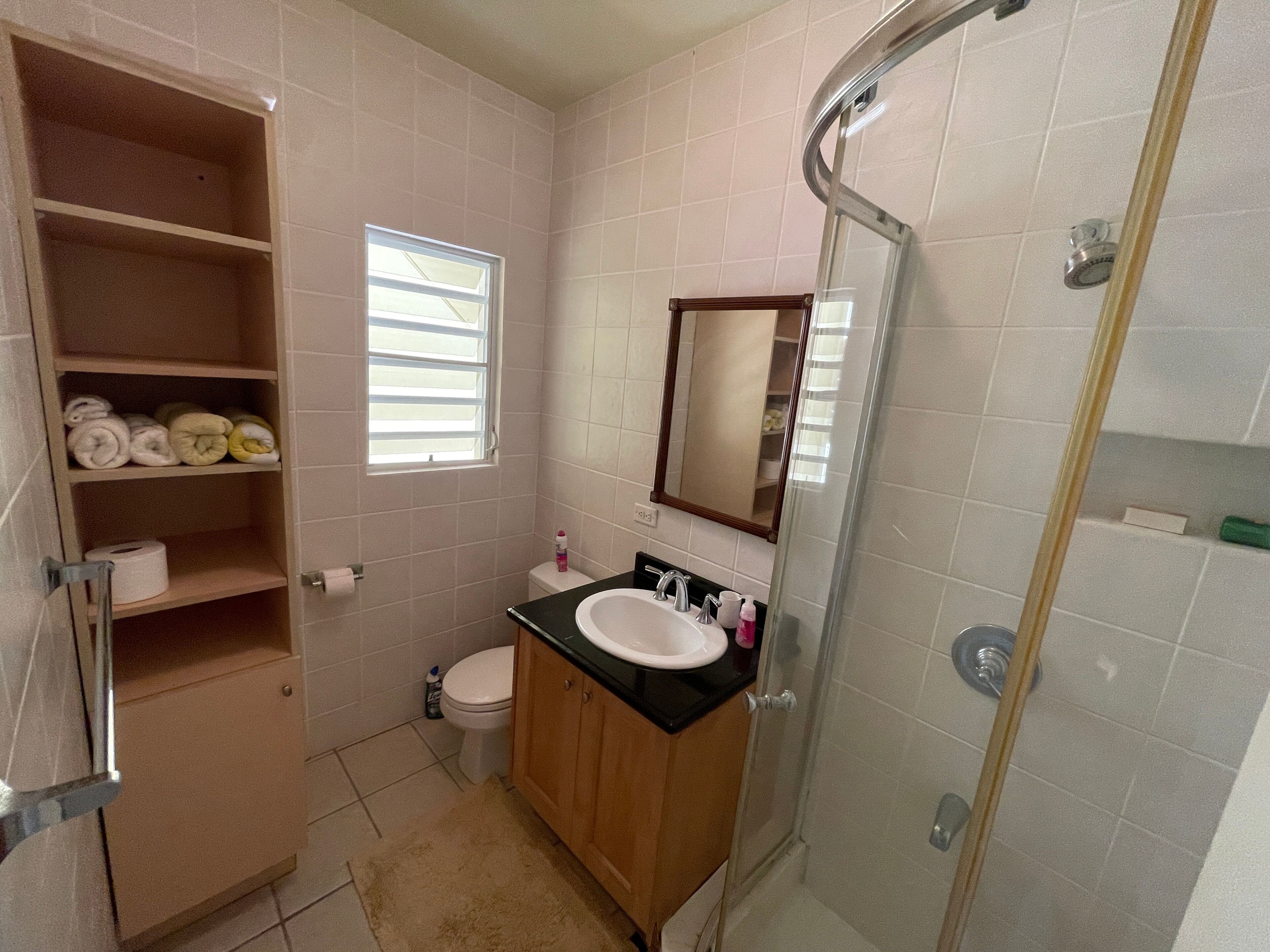 A bathroom with a toilet, sink, mirror, shower, and shelves with towels.