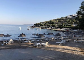 Una playa con tumbonas y sombrillas, barcos en el agua y una colina con edificios.