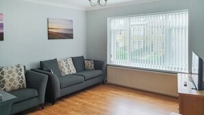 A living room with a grey sofa, patterned cushions, a wooden floor, a radiator, and a window with blinds.