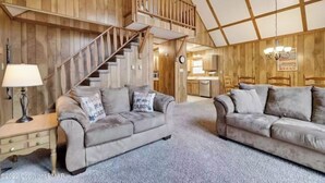 A cozy cabin interior with a wooden staircase, two grey sofas, a small desk, and a kitchen area.