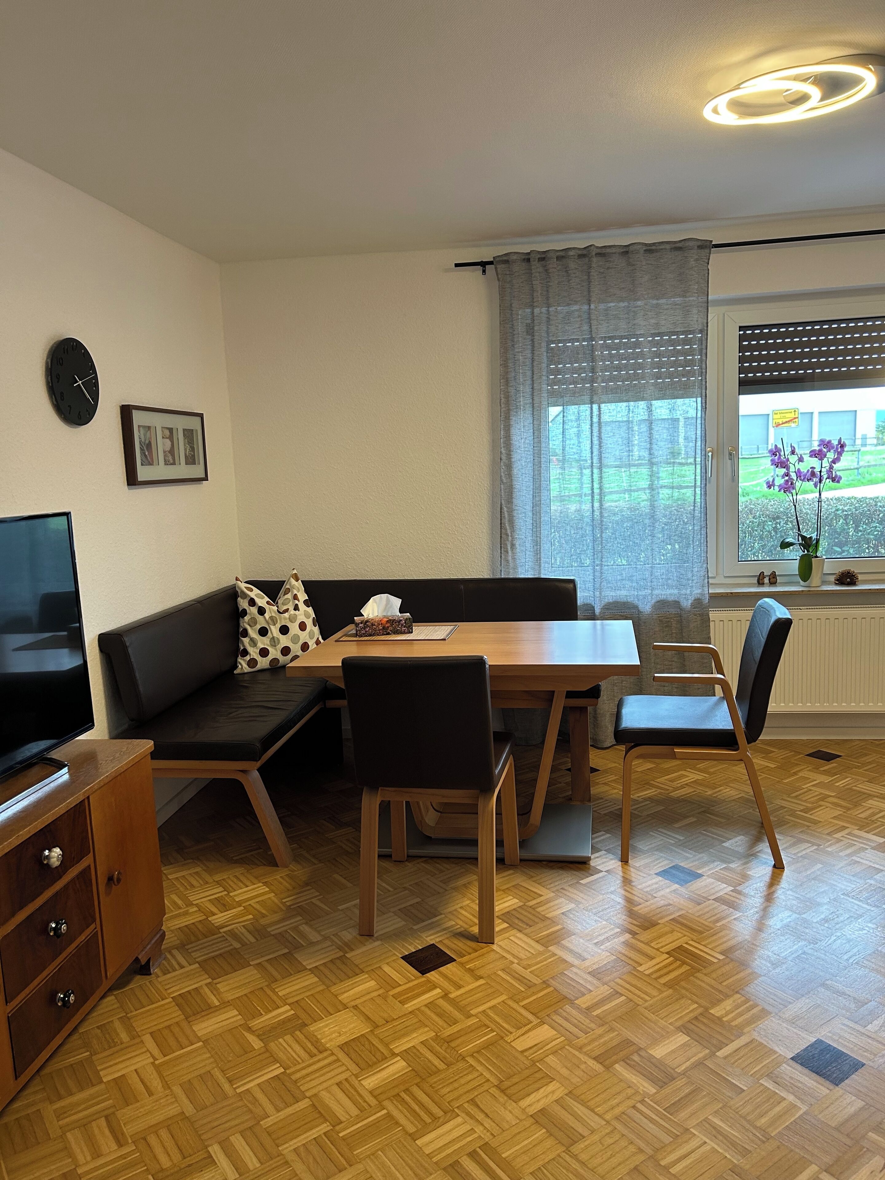 A modern living room with a wooden herringbone floor, a black leather sofa, a wooden dining table with chairs, a television, and a window with a view of greenery.