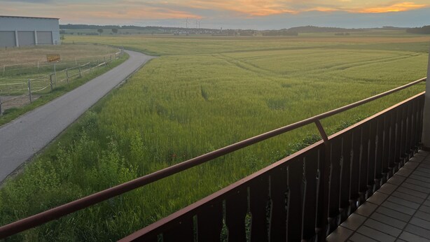 Ländliche Landschaft mit einer kurvenreichen Straße durch Felder, einem Gebäude links und einem Sonnenuntergang in der Ferne.