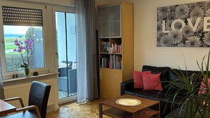 Ein Wohnzimmer mit Holzparkett-Boden, einer schwarzen Ledersofa, einem Couchtisch, einem Bücherregal und einem großen Fenster mit Blick auf Grünflächen.