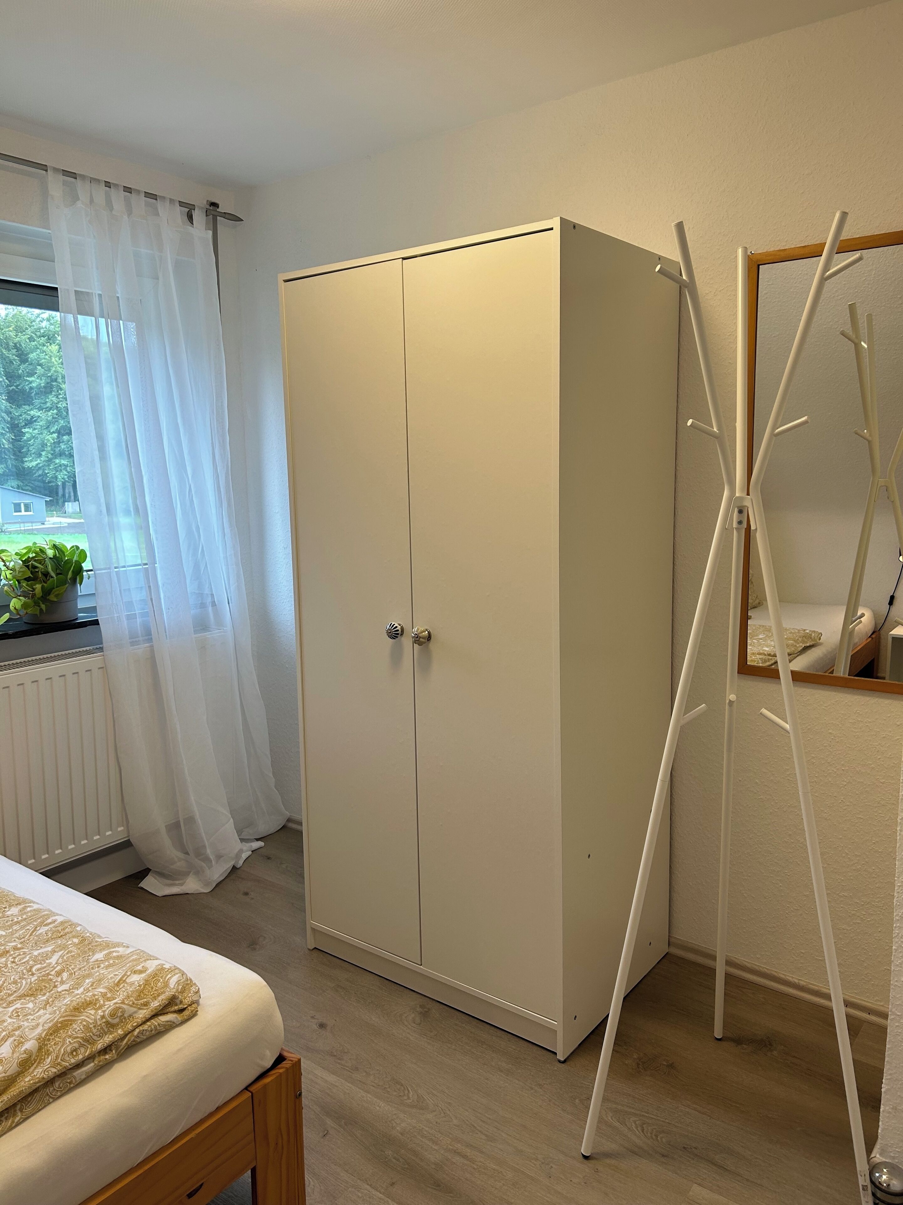 Et soveværelse med seng, skab, radiator og et vindue med let gardin.