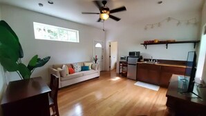 Amplia sala de estar con piso de madera, ventilador de techo, sofá, mesa de comedor y área de cocina con refrigerador y televisor.