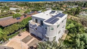 Una casa moderna de dos pisos con paneles solares en el techo, rodeada de abundante vegetación y vista al océano.