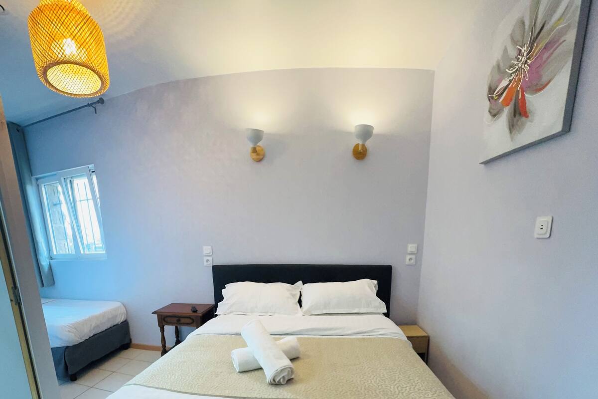 A neatly made bed with white linens, a bedside table with a lamp, and a window with blinds.