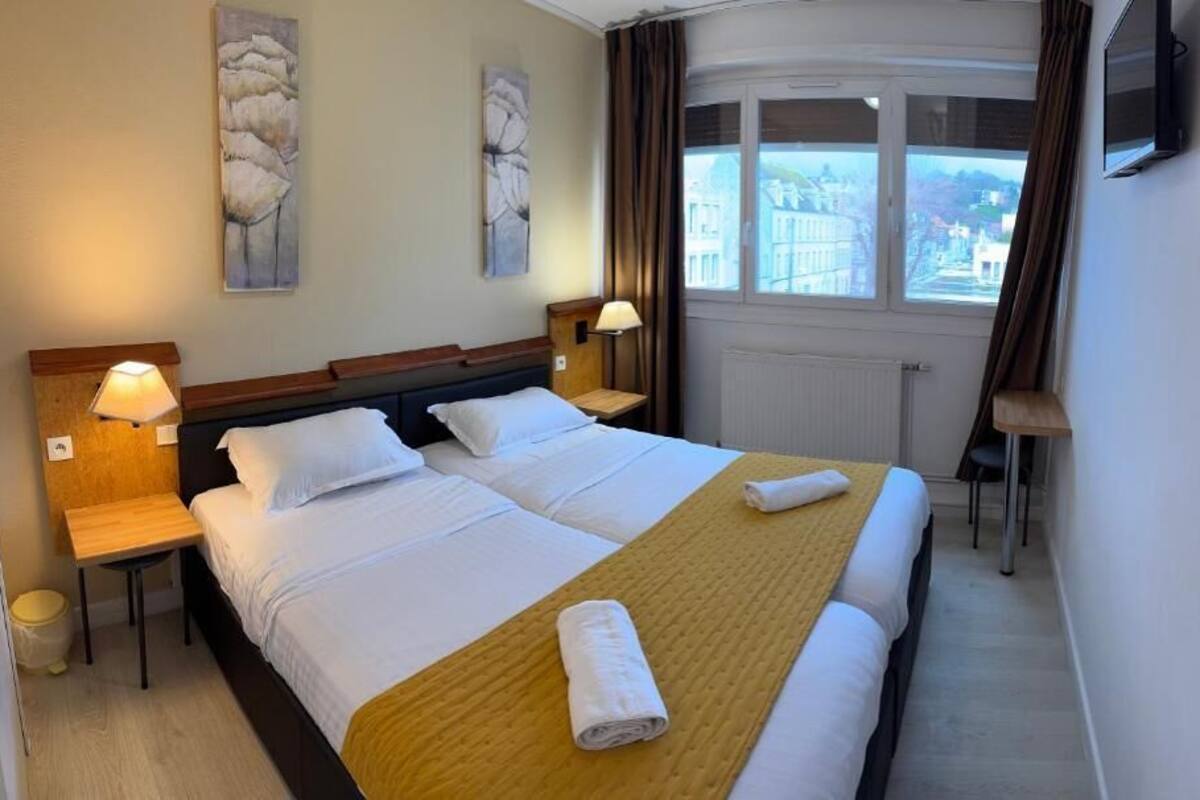 A hotel room with a double bed, two bedside tables with lamps, a view of the city through a window, and artwork on the walls.