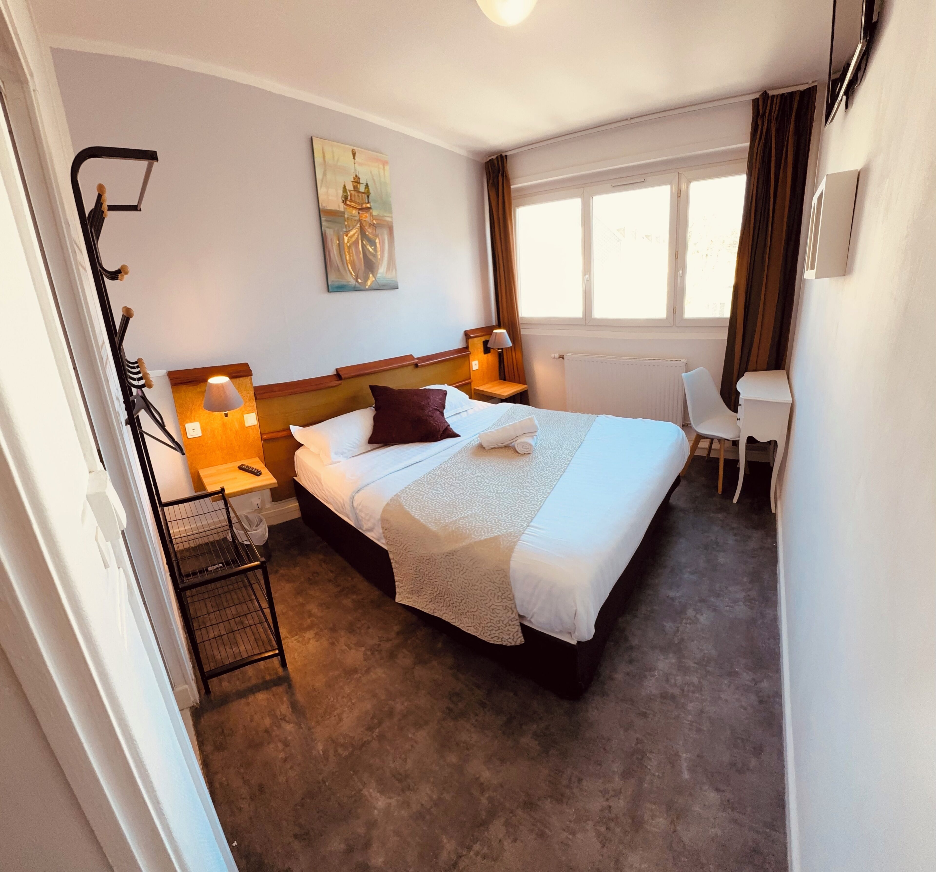 Habitación de hotel con cama, lámparas de noche, escritorio, silla, radiador y ventana con cortinas.