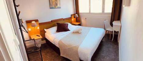 A hotel room with a bed, bedside lamps, a desk, a chair, a radiator, and a window with curtains.