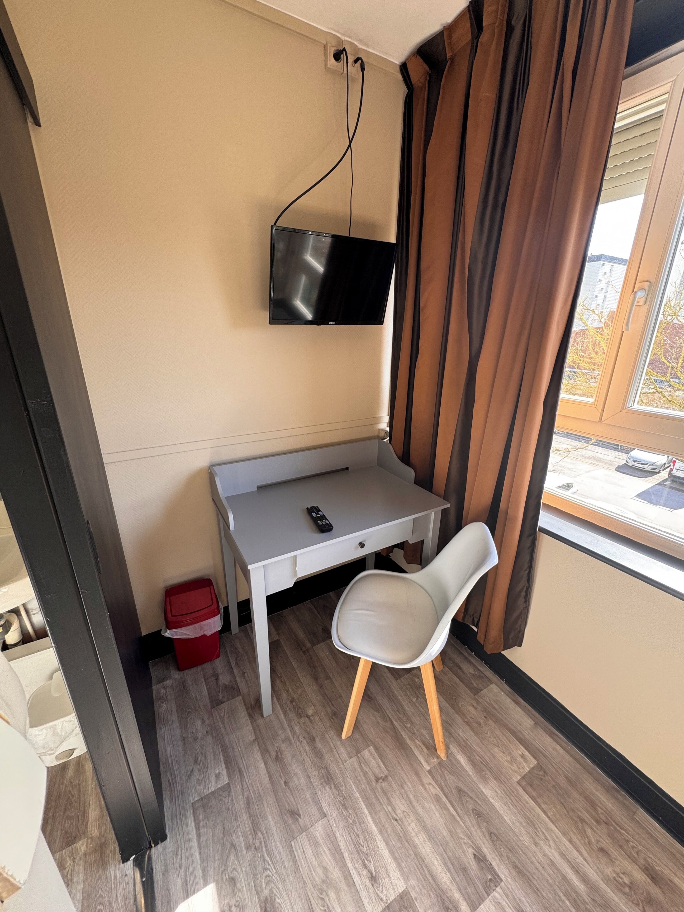 Une petite pièce comprenant un bureau, une chaise, un téléviseur fixé au mur et une fenêtre avec des rideaux.