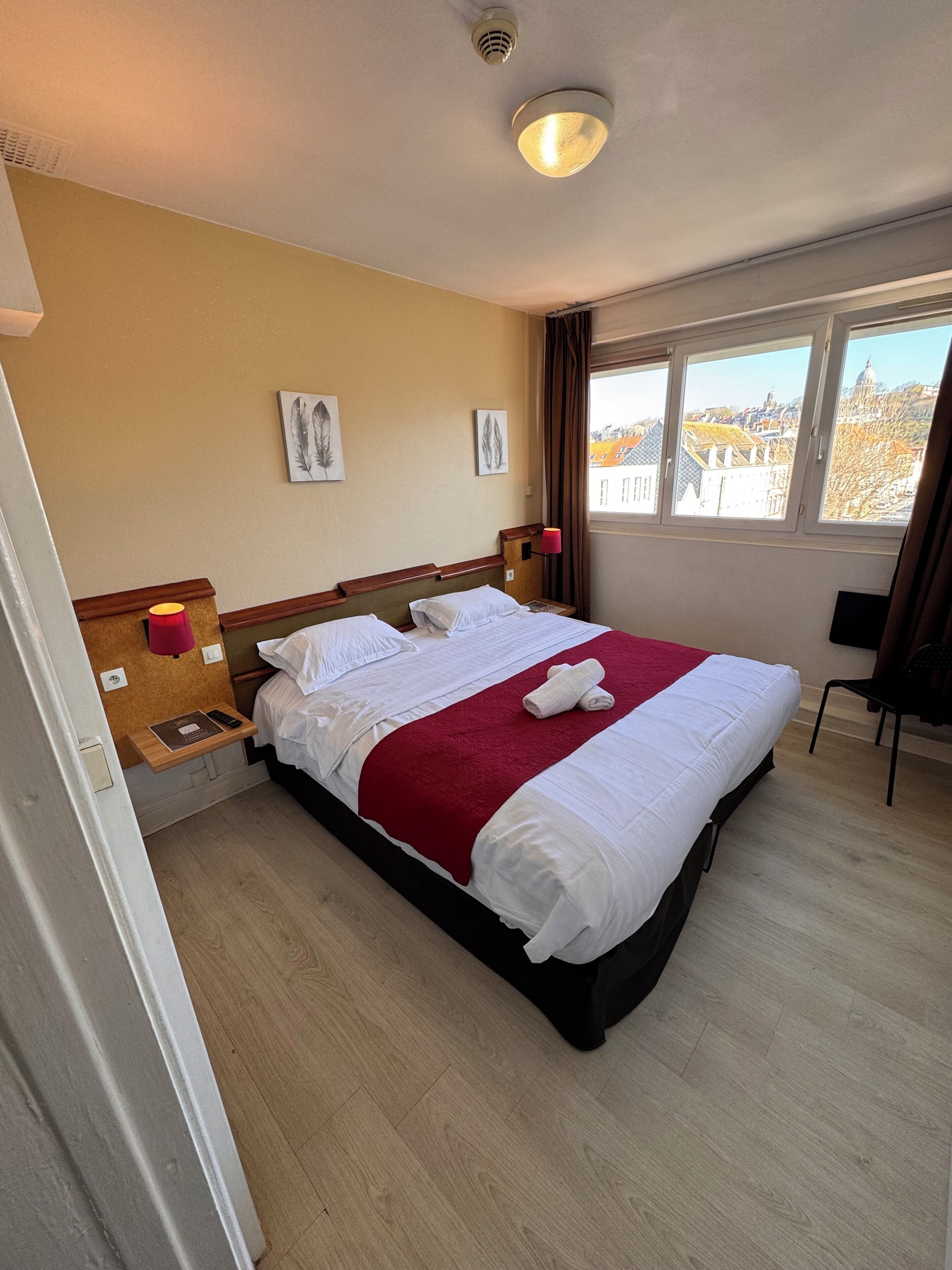 A hotel room with a bed, a bedside table, a window with a view, and a ceiling light.