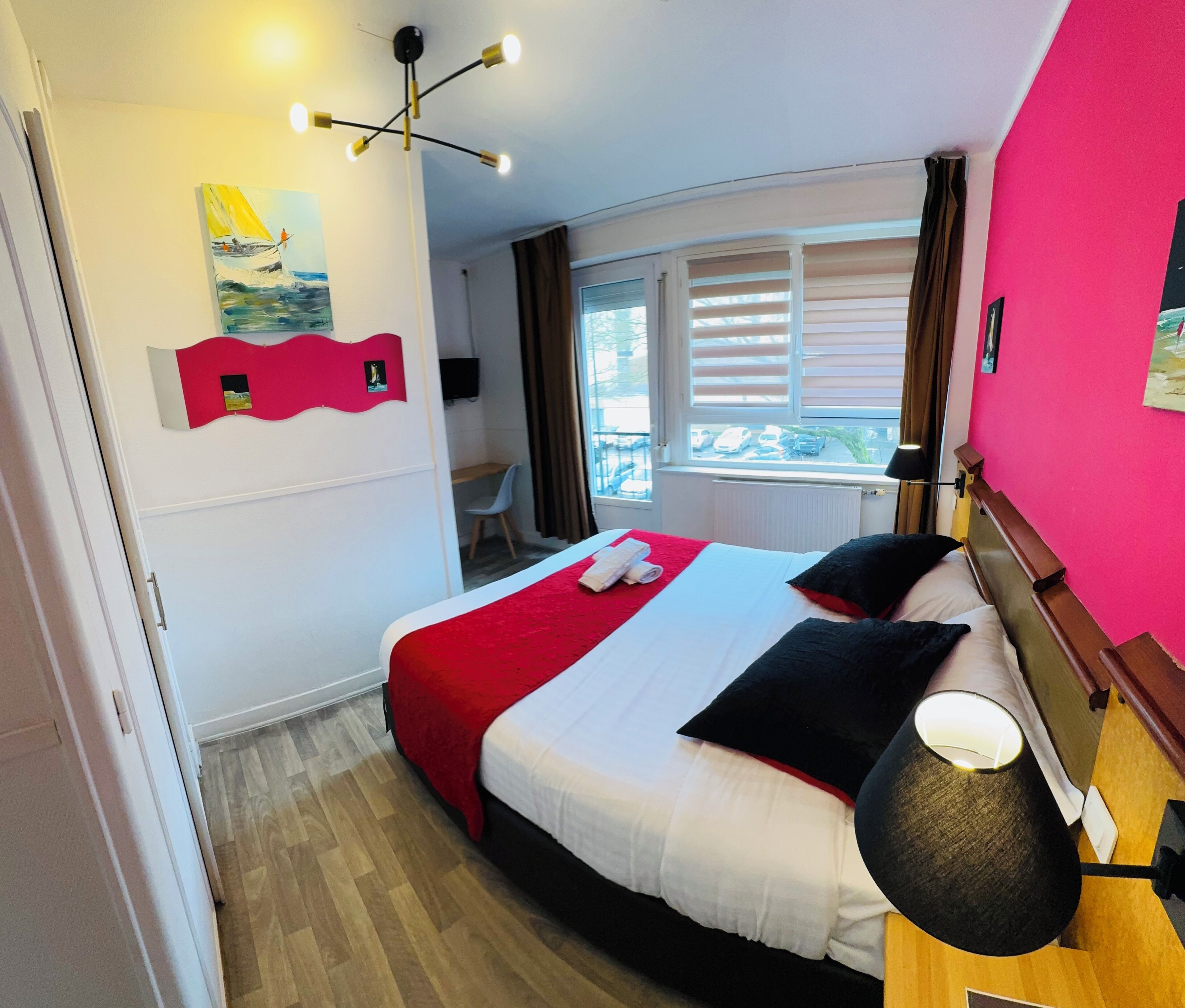 A hotel room with a bed, a desk, a chair, a window with blinds, a ceiling light, and artwork on the walls.