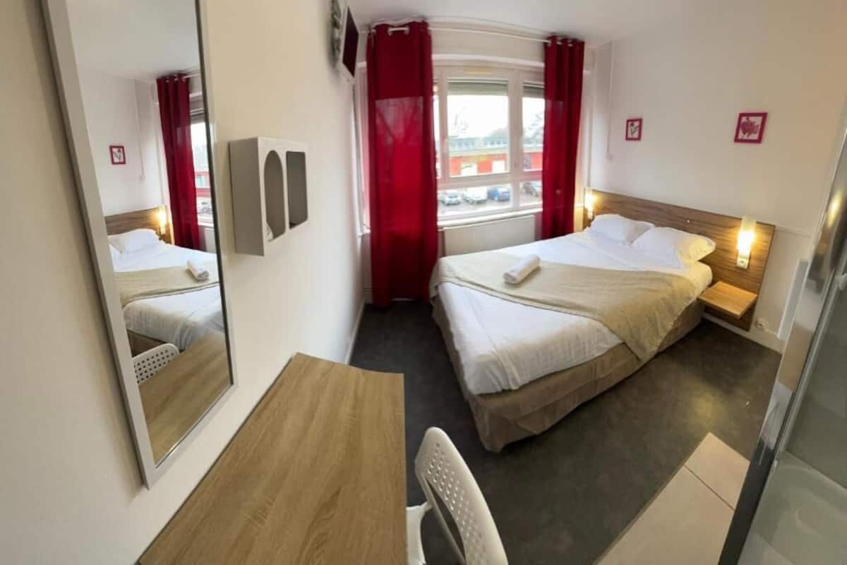 A hotel room with two beds, a desk, a chair, a mirror, and a window with red curtains.