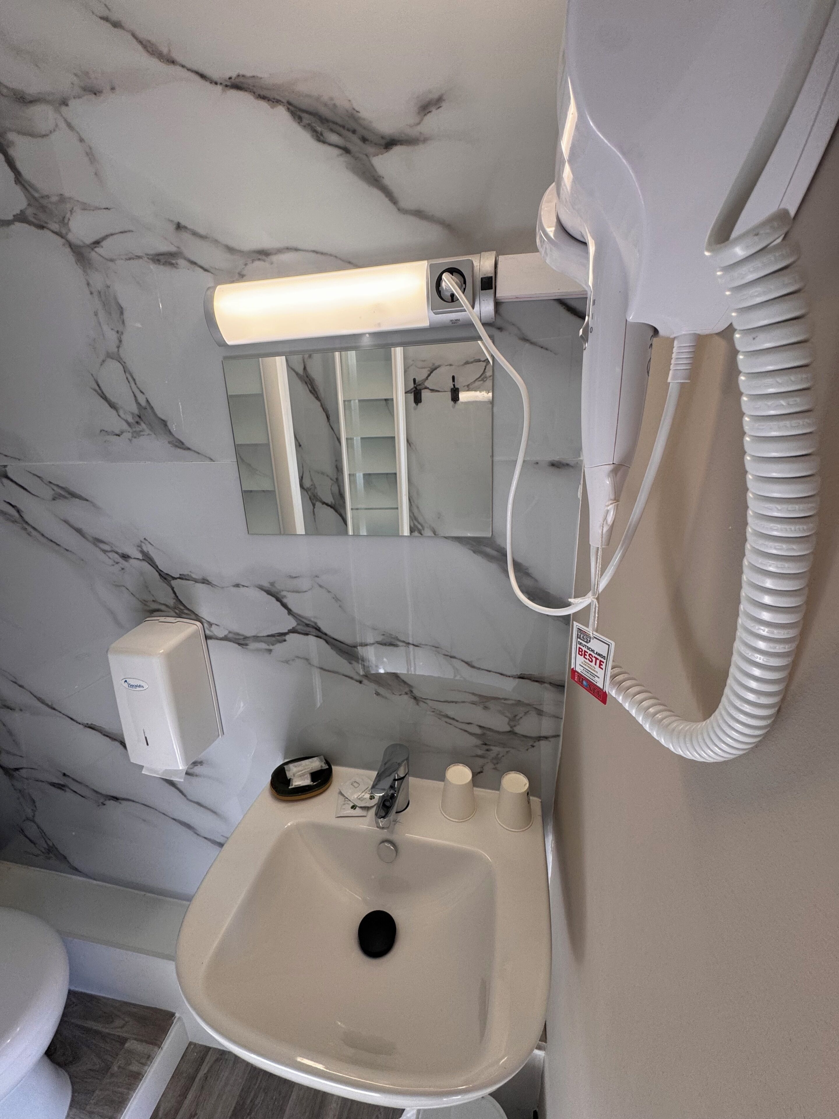 Un baño con pared de mármol, un lavamanos, un espejo, un secamanos y un inodoro.