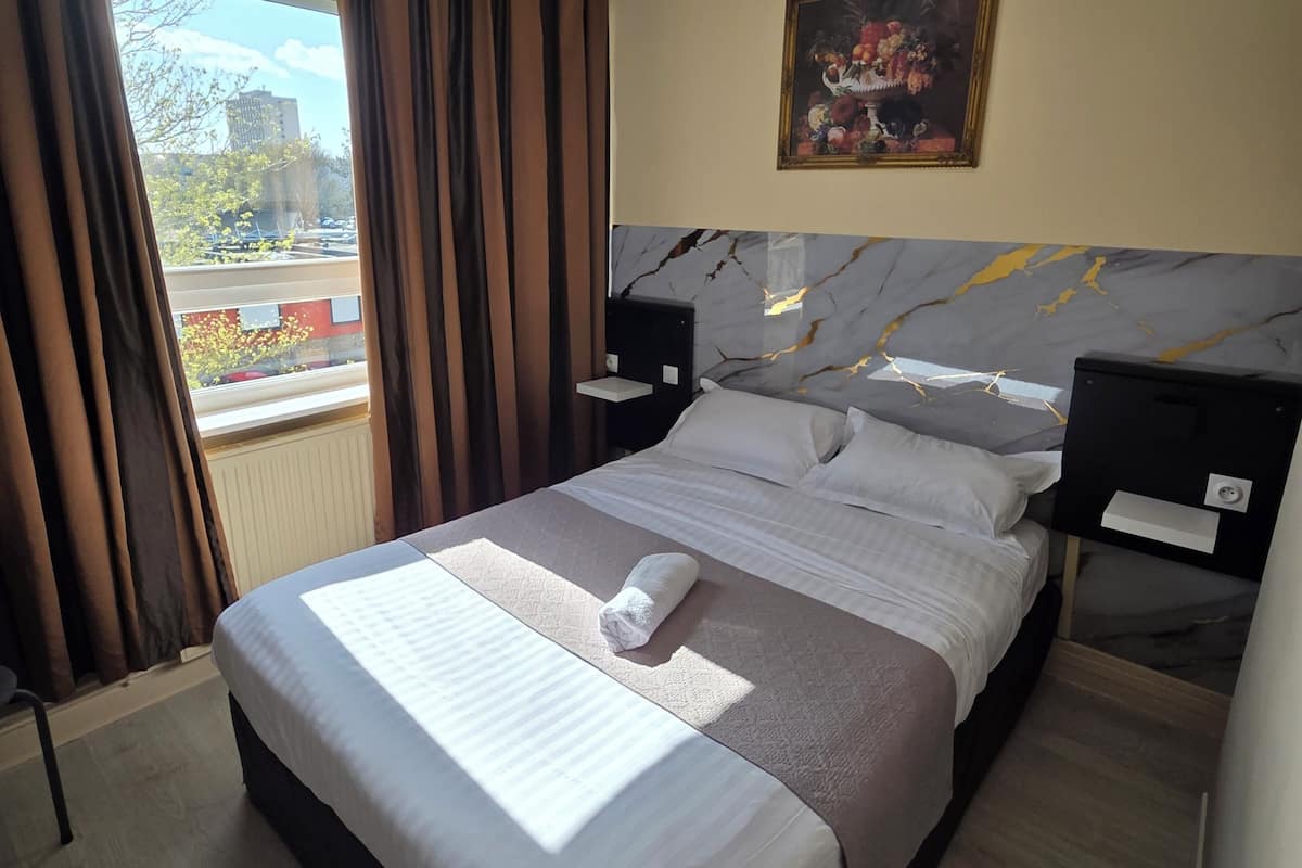 A hotel room with a bed, a nightstand, a painting, and a window with curtains.