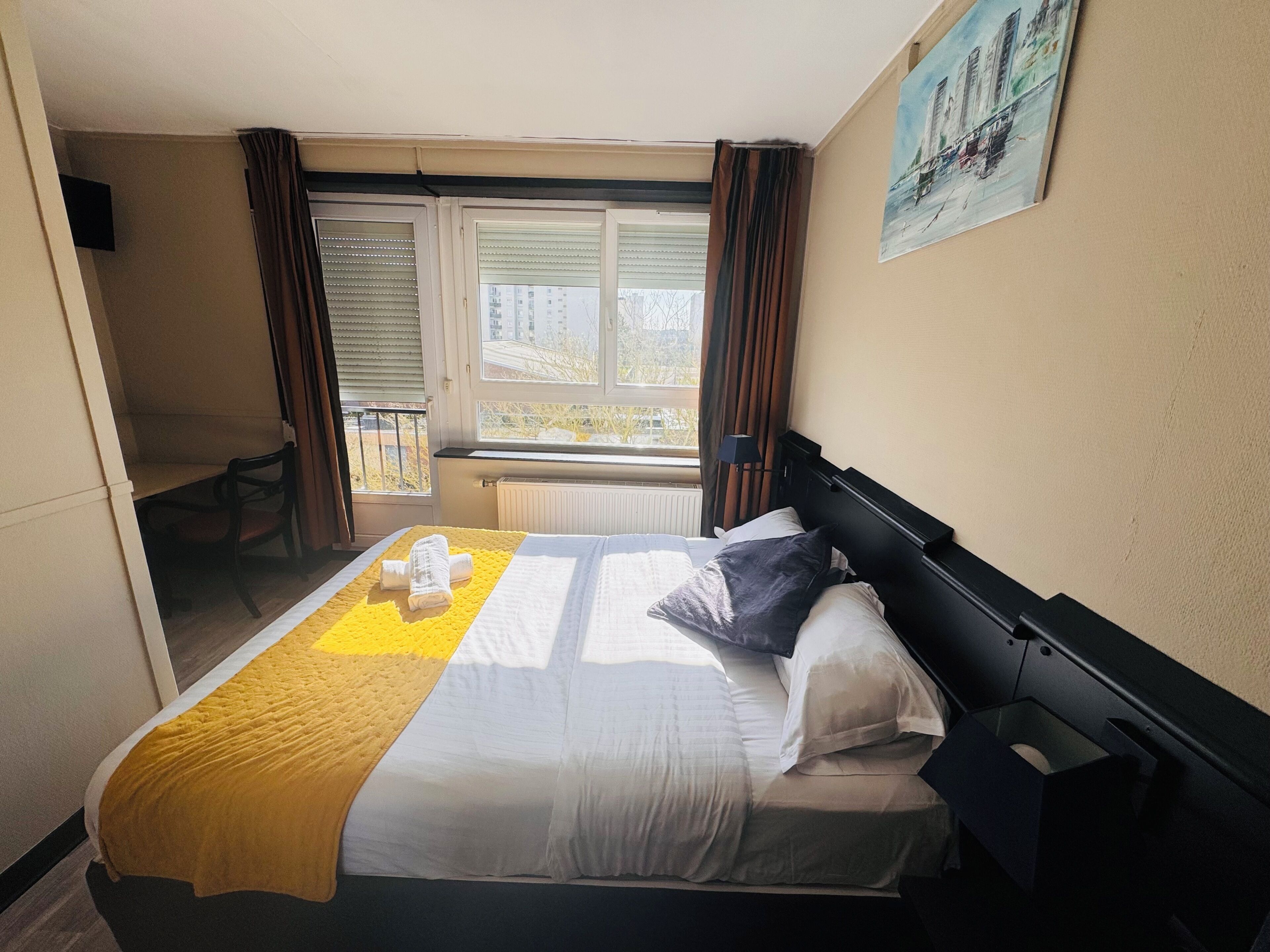 A hotel room with a bed, a window with curtains, a painting on the wall, and a chair by the window.