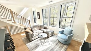 Un salon moderne avec un canapé d’angle, une table basse ronde et un fauteuil bleu. Un escalier avec des rampes en bois mène à un niveau supérieur.