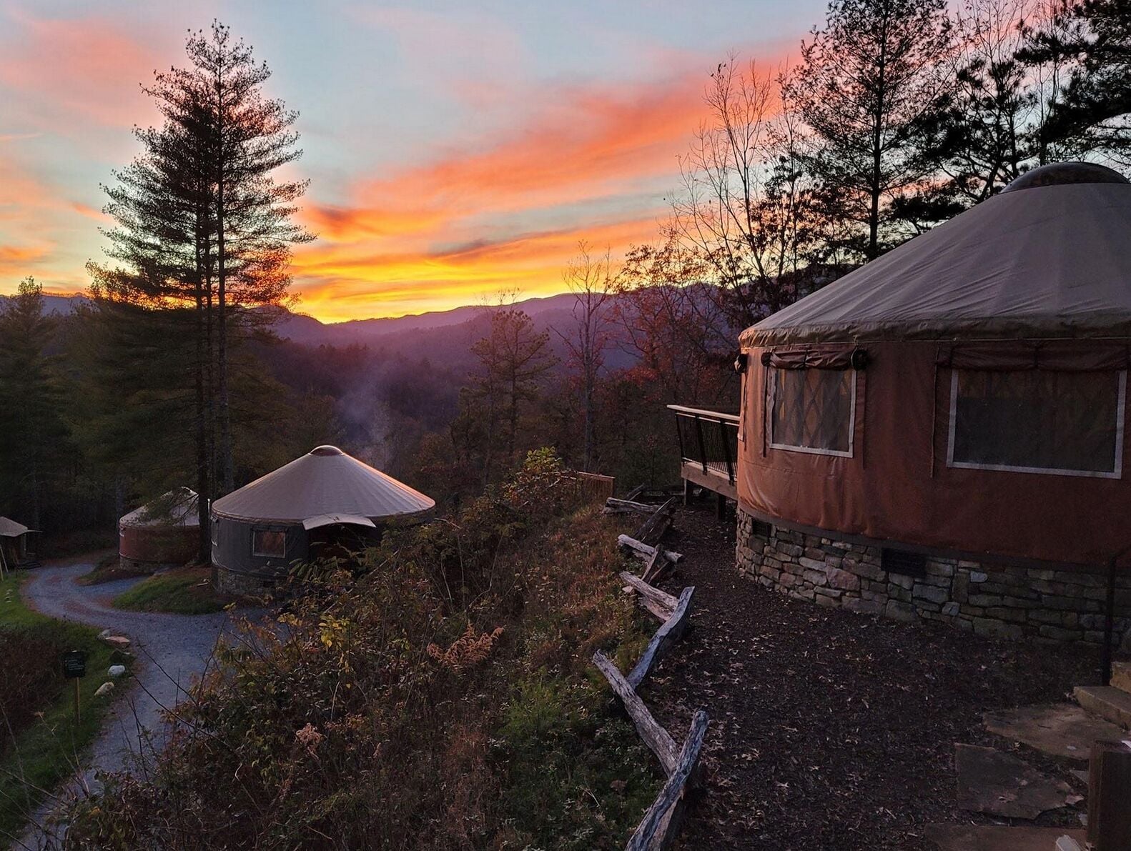 A rustic yurt-style tent with a stone foundation, surrounded by a forested landscape and a sunset in the background.