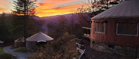 A rustic yurt-style tent with a stone foundation, surrounded by a forested landscape and a sunset in the background.