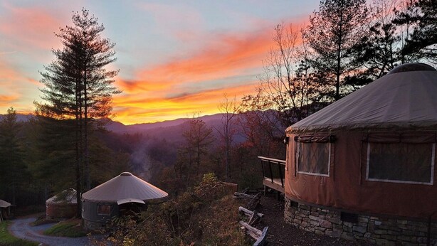 En rustik teltkonstruktion i yurts stil med stensokkel, omgivet af et skovlandskab og en solnedgang i baggrunden.
